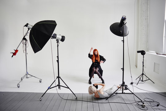 Girl photographer lies on the floor and shoots of fashion model posing on chair on white background in Studio