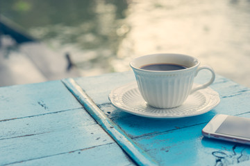 Cup of hot coffee with smartphone on old wooden table and view of sunrise in the river background.Vintage tone with copy space.Vacation concept