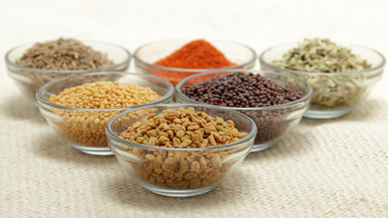 Different types of Indian spices in glass bowl, focus on Fenugreek seeds on jute mat background. Front view.