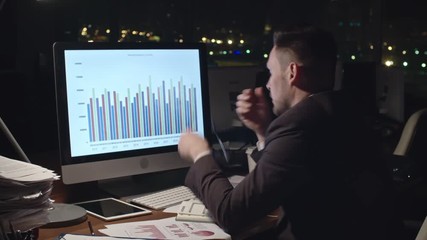 Medium shot of exhausted businessman analyzing financial statistics on computer screen, taking off his glasses, trying to concentrate and drinking coffee in dark office late at night - Powered by Adobe