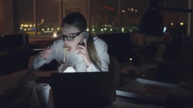 Young businesswoman sitting at desk in dark office, talking on mobile phone and eating noodles from box with sticks, open laptop in front of her, medium shot on Sony NEX700 + Odyssey 7Q