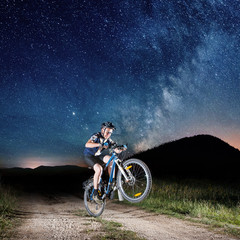 Courageous biker balancing on one wheel with his mountain bicycle in the night. Milky way shines above. Square photo