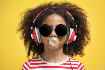 Afro-American little girl with headphones and sunglasses chewing gum on yellow background
