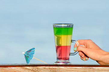 Female hand holding a glass with colorful cocktail on the background of the sea, umbrella for cocktails
