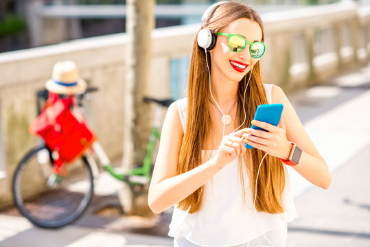 Young smiling woman listening to the music with headphones and smart phone standing on the street with bicycle on the background at the sunny weather