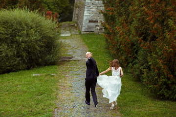 Fototapeta premium Groom waves with his hand while he runs with bride along the pat