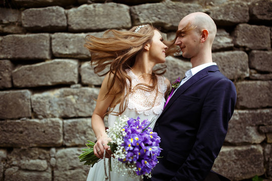 Bride touches groom's face with her hair standing behind a stone