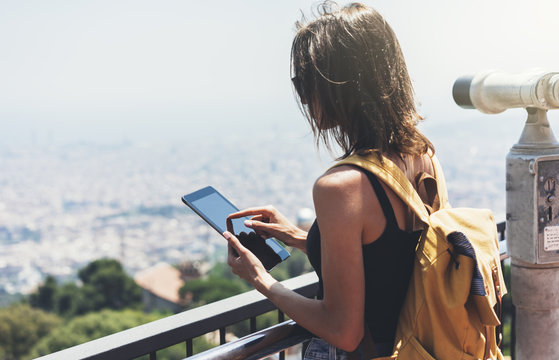 Hipster young girl with bright backpack planning travel plan. View from the back of the tourist traveler on background panoramic view Barcelona, coin operated binoculars. Female hands using tablet