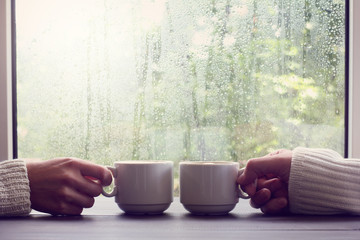 comfortable coffee break for two people/ two white cups in hands on a background of wet window with...