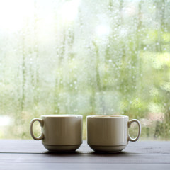 warming coffee in a cozy place/ pair of white cups are on the table against the background of wet window with raindrops 