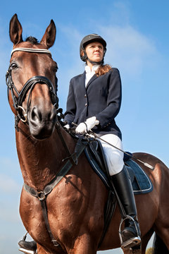 Portrait of serious beautiful young girl jockey in uniform sitting on a horse against blue sky and yellow field and looking forward on a sunny day. Equestrian sport - dressage.