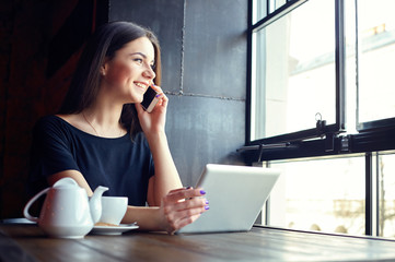 Young attractive girl talking on mobile phone and smiling while sitting alone in coffee shop during free time and working on tablet computer. Happy female having rest in cafe. Lifestyle