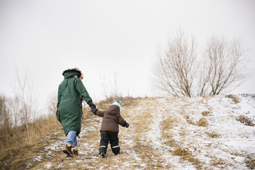 Small toddler boy and his mother walking the mountain trail on a winter day. Woman with little kid walking up the hill on a windy day. Boy following his mom on the valley trail.