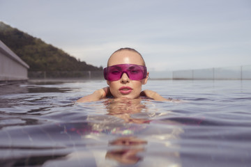 Face closeup of sexy young woman wearing purple sunglasses looking at camera in infinity rooftop swimming pool on a sunny day over blue sky and green trees landscape