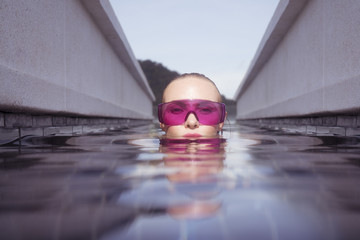 Fototapeta na wymiar Face closeup of sexy young woman wearing purple sunglasses looking at camera in infinity rooftop swimming pool on a sunny day over blue sky and green trees landscape