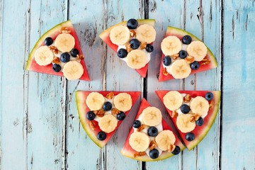 Sliced, juicy watermelon pizza with bananas, blueberries, nuts and yogurt, overhead view on rustic blue wood