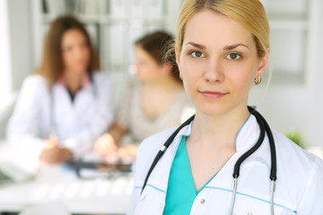 Young beautiful female doctor smiling  on the background with patient and his physician in hospital. High level and quality medical service concept.