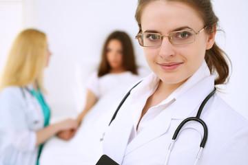 Fototapeta premium Young beautiful female doctor smiling on the background with patient and his physician in hospital. High level and quality medical service concept.