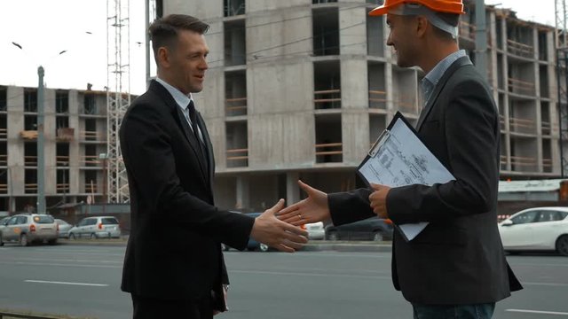 Slow motion. Two Young successful business people shaking hands. Architect in orange helmet with text pad, Customer in suit and tie. building under construction bg. Middle shot
