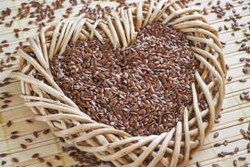 Flax seed in woven heart-shaped basket with flaxseed scattered around on bright wooden background.