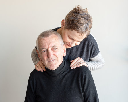 Older Man With Short Grey Hair And Black Top Seated And Older Woman With Short Hair Embracing Him From Behind With Hands On His Shoulders