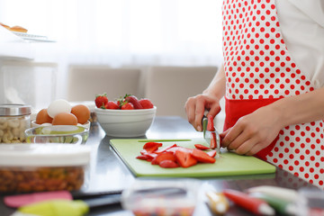Obraz na płótnie Canvas Young beautiful woman dressed in a white shirt and white red polka dot pinafore,engaged in the bright kitchen slicing red ripe strawberries to decorate cupcakes