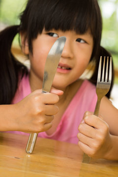 little asian girl waiting for dinner. Holding a knife and fork in the hand

