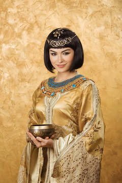 Beautiful woman like Egyptian Queen Cleopatra with cup on golden background