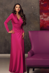 Fototapeta na wymiar Young beautiful female model with perfect shapes wearing long evening pink dress and standing near purple armchair. Model looking at you or in camera
