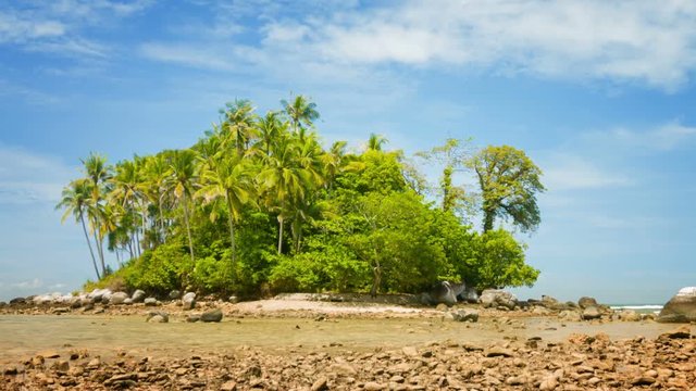 Video - Small tropical island at low tide, under a blue sky with cirus clouds. Lots of coconut palms and other trees. A rocky beach with boulders. Shallow tide-pool in the foreground