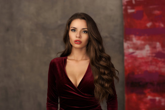 Young beautiful and pretty elegant woman in long cherry red evening dress posing against gray concrete wall. Portrait of smiling happy cheerful girl