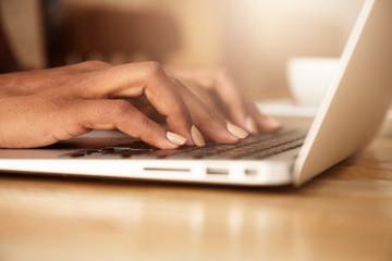 Cropped portrait of young female student learning on-line using generic laptop computer. Dark-skinned business woman sitting in front of open net-book, typing a message. Selective focus on fingers