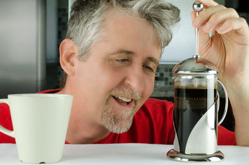 A happy sleepy man with messy morning hair is smiling as he uses his French press coffee maker which makes an exceptional caffeine brew