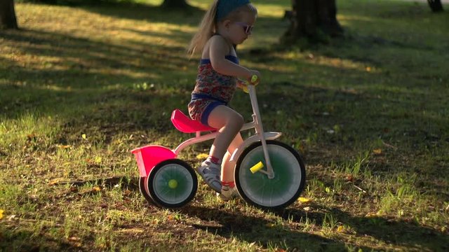 A little girl rides in the evening on a pink tricycle through the city Park