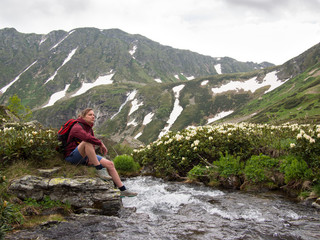 Young woman with backpack sitting on the bank of the river with flowers on the background of snowy mountains and clouds