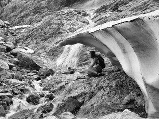 Monochrome image adult woman with backpack sitting on a rock under a block of ice on a background of a mountain with a waterfall