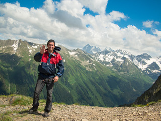 A young man in a jacket stands near the cliff and smiles on the background of the North Caucasus mountain range and cloudy sky