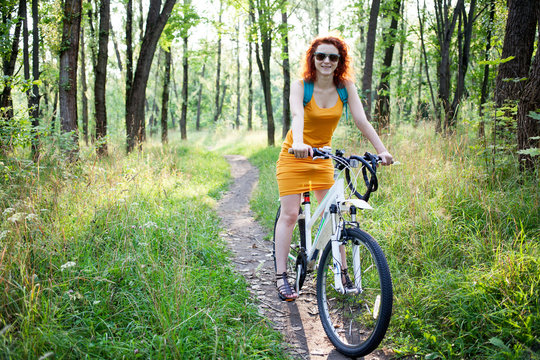Young ginger woman in sunglasses riding a bike on a path in the forest
