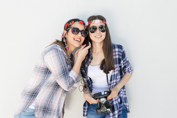 Two smiling young girls are standing in front of the white wall and listening to a music. One of them holing an old camera.