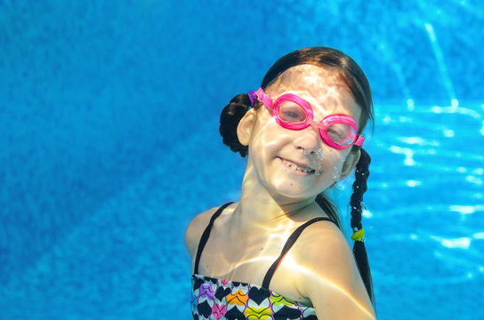 Child swims in pool under water, funny happy girl in goggles has fun and makes bubbles, kid sport on active family vacation
