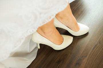 Close up of beautiful thin legs of young bride and bridal white dress over brown wood floor background. Woman putting on classic shoes heels for wedding ceremony.