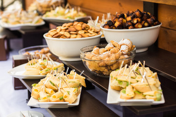 Wood brown tables full of different snacks for party or wedding celebration. White plates with cheese sandwiches and variety of sweet small cookies. Catering for wedding celebration in restaurant.