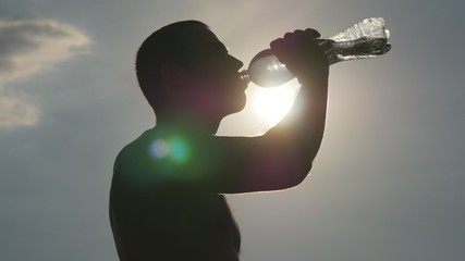 Young man drinking water from a plastic bottle in nature. Guy having water break at sunset. The Sun in the Background. Silhouette of male profile