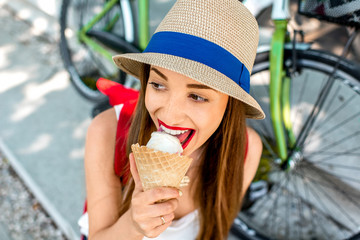 Young female tourist with backpack and hat enjoying ice cream sitting on the street near the bicycle in the city. Image with small depth of field and view from above