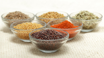 Different types of Indian spices in glass bowl, focus on brown mustard seeds on jute mat background. Front view.