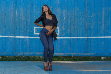 Young woman in top, shirt and leggins on high heels with modern makeup and hair dancing against blue painted wooden wall