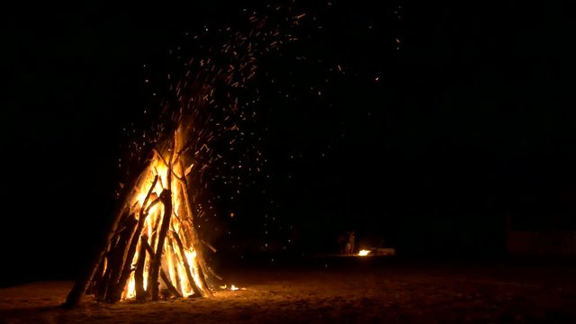 A huge bonfire on the sand and a small fire in the background at night. Lots of sparks from the burning logs. People stand near the fire and talk.