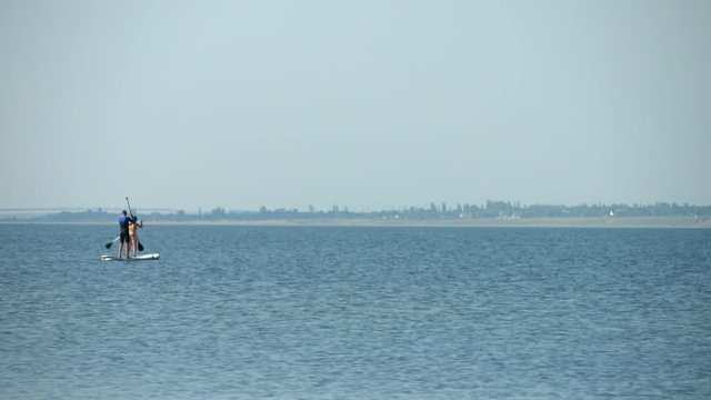 Young couple rides and floates on inflatable board for stand up paddling on the river. Quiet calm expanse of water in the lake. A man in a diving suit, girl in bikini swimsuit. Silhouettes of family