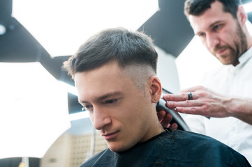 Fototapeta premium Professional styling. Close up side view of young man getting haircut by hairdresser with electric razor at barbershop