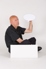 Älterer Mann sitzt vor weißem Schild und hält Sprechblase